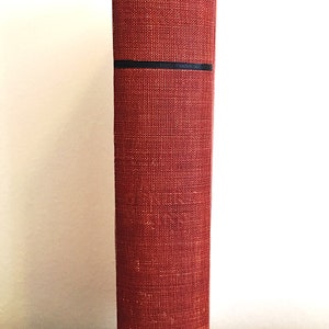 Może przedstawiać: Grzbiet czerwonej książki w twardej oprawie z czarnymi paskami. Książka stoi pionowo na szarej powierzchni.