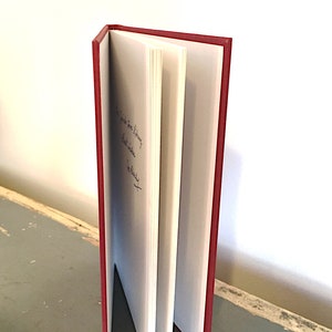 May include: A red hardcover book with a white spine and white pages. The book is open and the pages are fanned out. The book has a handwritten inscription on the inside cover.