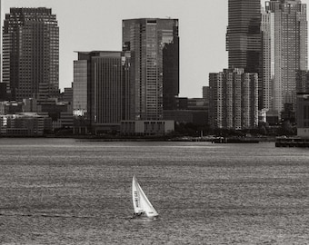 May include: A black and white photograph of a cityscape with a sailboat in the foreground. The sailboat has a white sail with the text "Mount Gay" printed on it. The cityscape is made up of tall buildings, some of which are partially obscured by clouds.