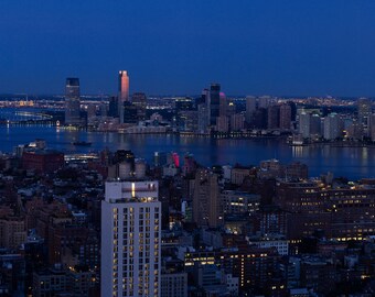 Pode incluir: Vista noturna de uma cidade a partir de um edifício alto, mostrando um grupo de arranha-céus à distância do outro lado de um corpo de água. Em primeiro plano, pode-se ver uma área urbana densa com edifícios de diferentes alturas.