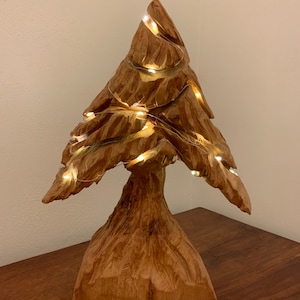 Può includere: Una scultura di albero di Natale in legno con luci avvolte intorno. L'albero è scolpito con un aspetto rustico e naturale e ha una base larga.