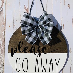May include: A round wooden sign with a black and white checkered bow. The sign is painted white on the bottom half and brown on the top half. The text "please go away" is written in black on the white half of the sign.