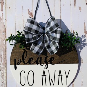May include: A round wooden door hanger with a black and white checkered bow, green leaves, and the text "please go away" in black lettering.