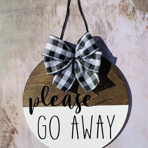 May include: A round wooden sign with a black and white checkered bow. The sign is painted white on the bottom half and brown on the top half. The text "please go away" is written in black on the white half of the sign.