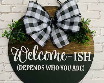 May include: A round wooden door hanger with a black and white checkered bow, green foliage, and the text "Welcome-ISH (DEPENDS WHO YOU ARE)".