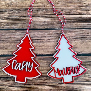 K&ouml;nnte beinhalten: Zwei rote und wei&szlig;e Weihnachtsbaumschmuckst&uuml;cke mit den Namen "Carly" und "Maverick" darauf.