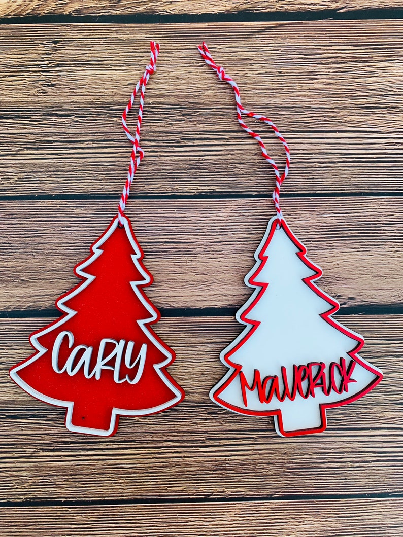 K&ouml;nnte beinhalten: Zwei rote und wei&szlig;e Weihnachtsbaumschmuckst&uuml;cke mit den Namen "Carly" und "Maverick" darauf.