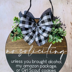 Puede incluir: Un letrero redondo de madera con un lazo a cuadros blanco y negro. El letrero dice "no soliciting unless you brought alcohol, my amazon package, or Girl Scout cookies."