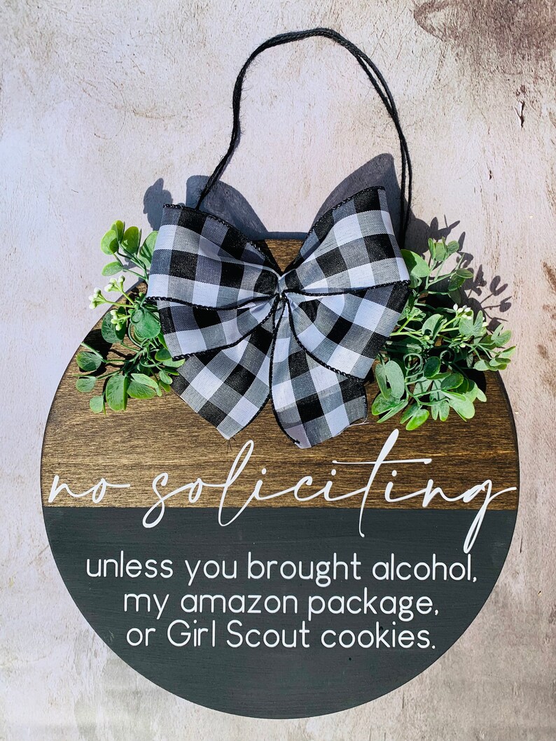 Puede incluir: Un letrero redondo de madera con un lazo a cuadros blanco y negro. El letrero dice "no soliciting unless you brought alcohol, my amazon package, or Girl Scout cookies."
