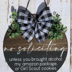 Puede incluir: Un letrero redondo de madera con un lazo a cuadros blanco y negro y vegetaci&oacute;n. El letrero dice "no soliciting unless you brought alcohol, my amazon package, or Girl Scout cookies."