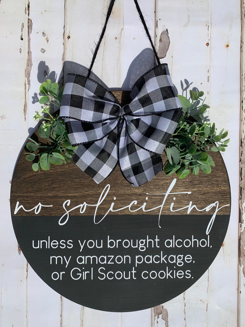 Puede incluir: Un letrero redondo de madera con un lazo a cuadros blanco y negro y vegetaci&oacute;n. El letrero dice "no soliciting unless you brought alcohol, my amazon package, or Girl Scout cookies."