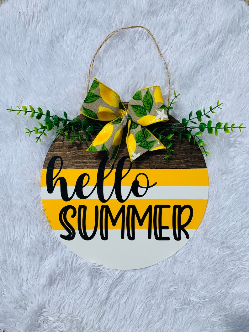 May include: A round wooden sign with a yellow, white, and brown striped design. The sign says "hello summer" in black lettering. A lemon-themed bow is attached to the top of the sign.