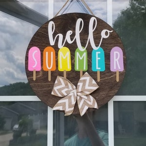 May include: A round wooden sign with the text "hello summer" in white lettering. The sign is decorated with six colorful popsicle shapes, each with a different color: pink, orange, yellow, green, blue, and purple. A brown and white bow is attached to the bottom of the sign.