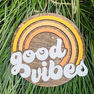 May include: A round wooden sign with a brown background and a white inscription that reads "good vibes". The sign is decorated with a multi-colored rainbow design.
