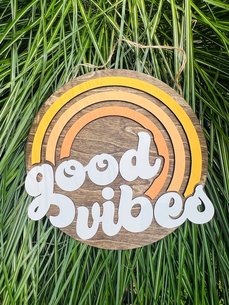 May include: A round wooden sign with a brown background and a white inscription that reads "good vibes". The sign is decorated with a multi-colored rainbow design.