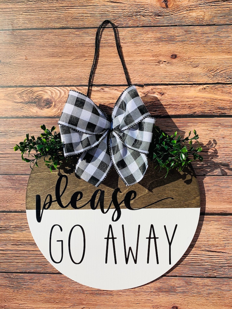May include: A round wooden sign with a white and brown background. The sign says "please go away" in black lettering. The sign is decorated with a black and white checkered bow and green foliage.