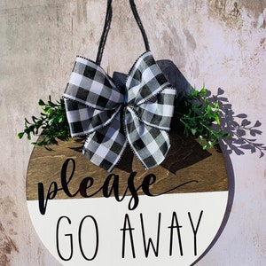 May include: A round wooden sign with a black and white checkered bow, green foliage, and the text "please go away" in black lettering.