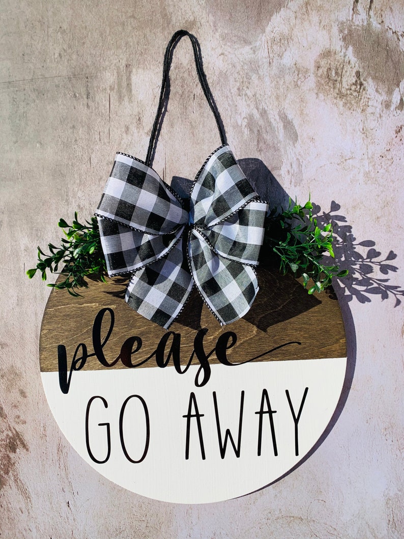 May include: A round wooden sign with a black and white checkered bow, green foliage, and the text "please go away" in black lettering.