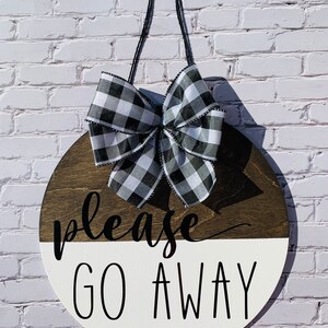 May include: A round wooden sign with a black and white buffalo check bow. The sign is painted white on the bottom half and brown on the top half. The text "please go away" is written in black on the sign.