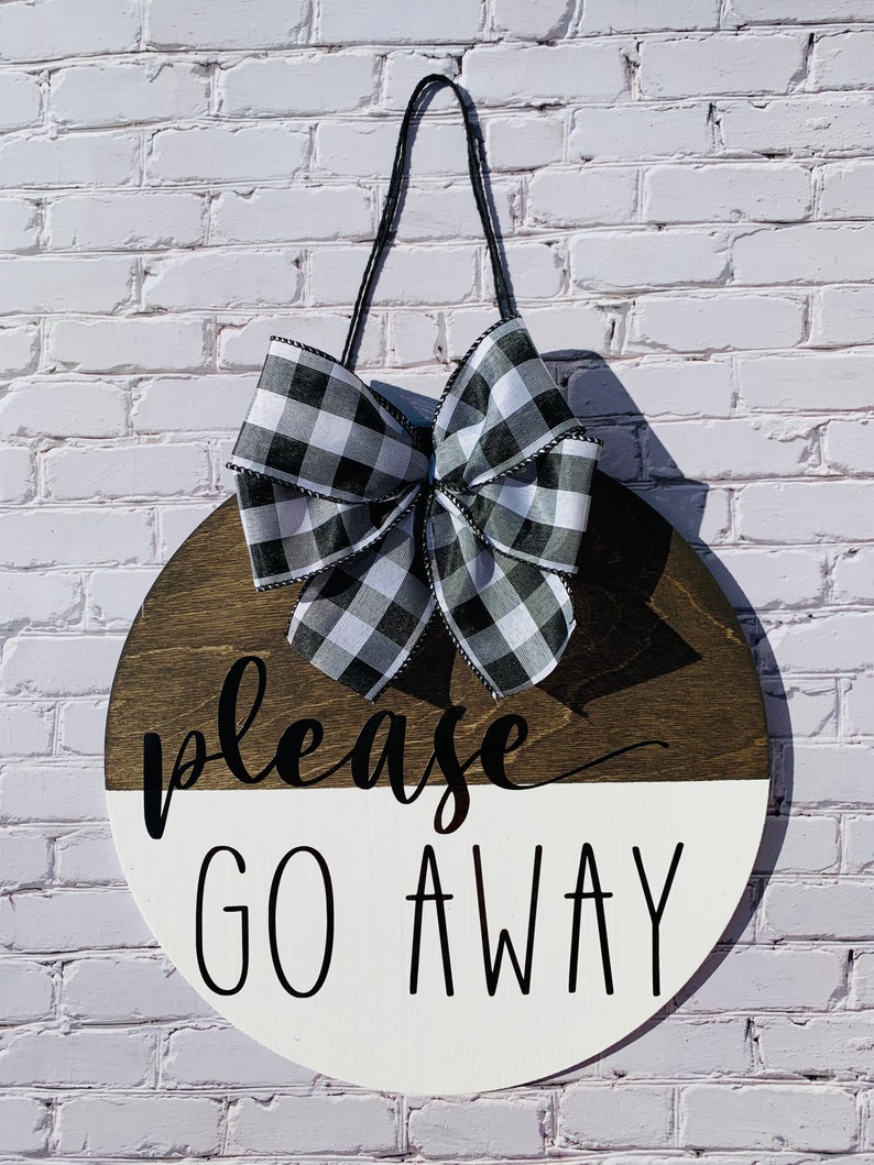 May include: A round wooden sign with a black and white buffalo check bow. The sign is painted white on the bottom half and brown on the top half. The text "please go away" is written in black on the sign.
