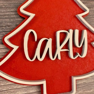 K&ouml;nnte beinhalten: Ein roter Weihnachtsbaum-Anh&auml;nger aus Holz mit wei&szlig;em Rand und dem Namen "Carly" in wei&szlig;er Kursivschrift.