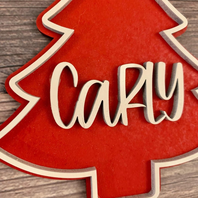 K&ouml;nnte beinhalten: Ein roter Weihnachtsbaum-Anh&auml;nger aus Holz mit wei&szlig;em Rand und dem Namen "Carly" in wei&szlig;er Kursivschrift.
