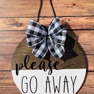 May include: A round wooden sign with a black and white checkered bow. The sign is painted white on the bottom half and brown on the top half. The text "please go away" is written in black on the sign.