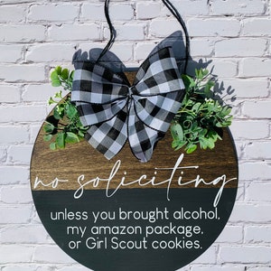 Puede incluir: Un letrero redondo de madera con un lazo a cuadros blanco y negro y vegetaci&oacute;n. El letrero dice "no soliciting unless you brought alcohol, my amazon package, or Girl Scout cookies."
