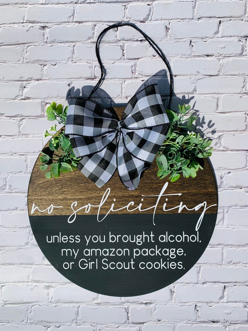 Puede incluir: Un letrero redondo de madera con un lazo a cuadros blanco y negro y vegetaci&oacute;n. El letrero dice "no soliciting unless you brought alcohol, my amazon package, or Girl Scout cookies."