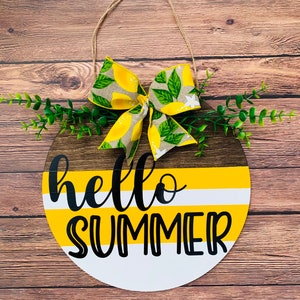 May include: A round wooden sign with a yellow and white striped background and the text "hello summer" in black. The sign is decorated with a lemon-themed bow and green foliage.