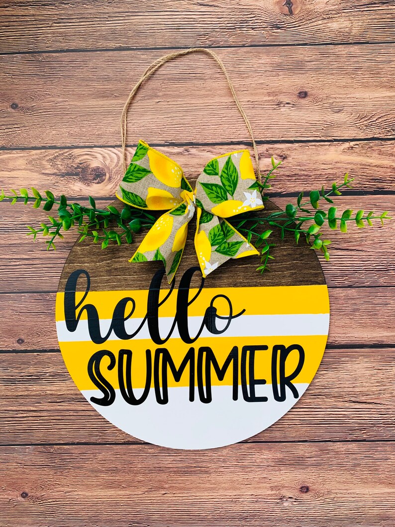 May include: A round wooden sign with a yellow and white striped background and the text "hello summer" in black. The sign is decorated with a lemon-themed bow and green foliage.
