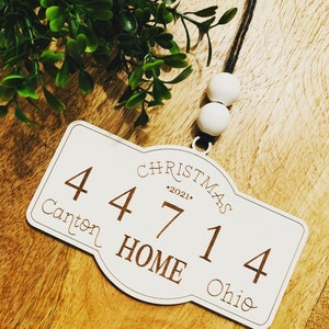 Op de afbeelding: Witte houten kerstversiering met de tekst "Christmas 2021" en "Home" in een cursief schrift. De versiering heeft ook de nummers "44714" en de tekst "Canton Ohio".