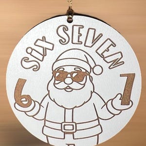 May include: A circular white Christmas decoration featuring the words "SIX SEVEN" and a cartoon Santa Claus wearing sunglasses. The Santa holds the numbers 6 and 7. A gold string is attached for hanging.