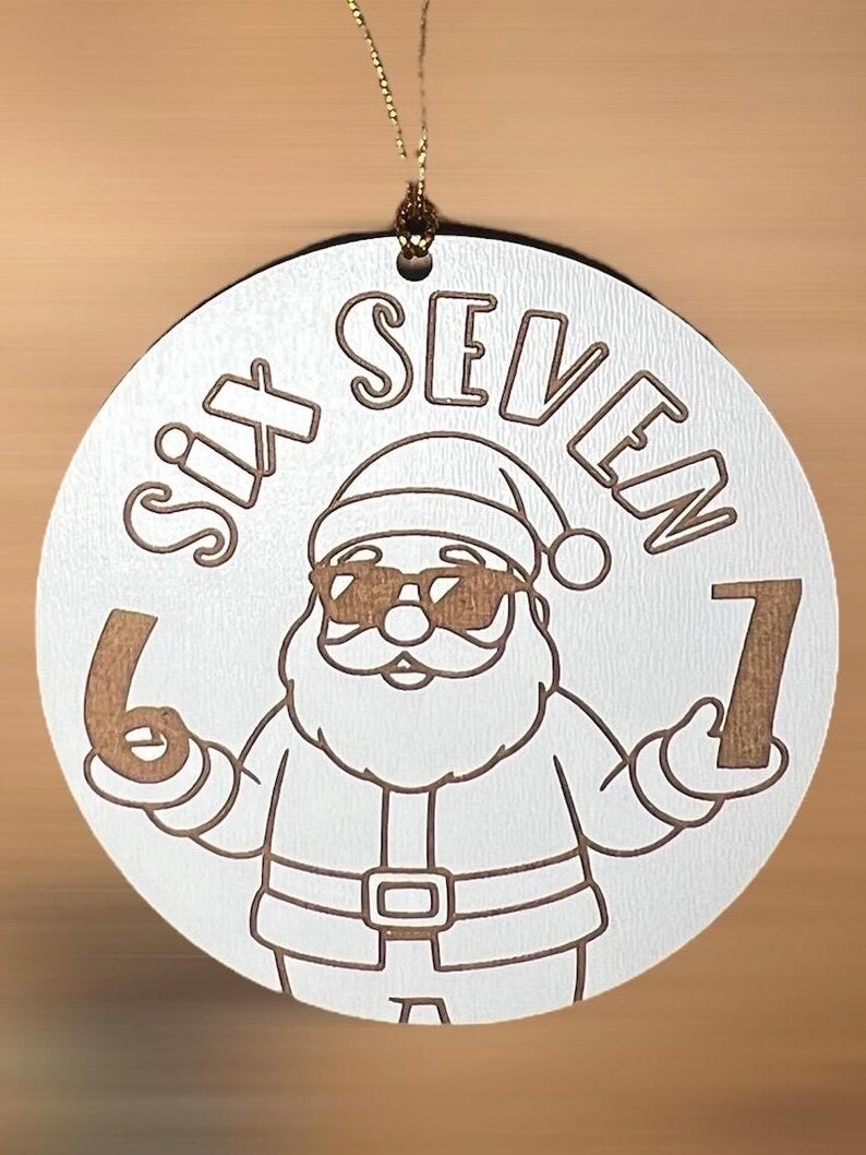 May include: A circular white Christmas decoration featuring the words "SIX SEVEN" and a cartoon Santa Claus wearing sunglasses. The Santa holds the numbers 6 and 7. A gold string is attached for hanging.