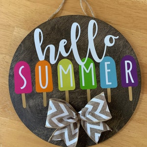 May include: A round wooden sign with the words "hello summer" in white script. The word "summer" is spelled out with colorful popsicle-shaped letters. A brown and white chevron bow is tied at the bottom of the sign.