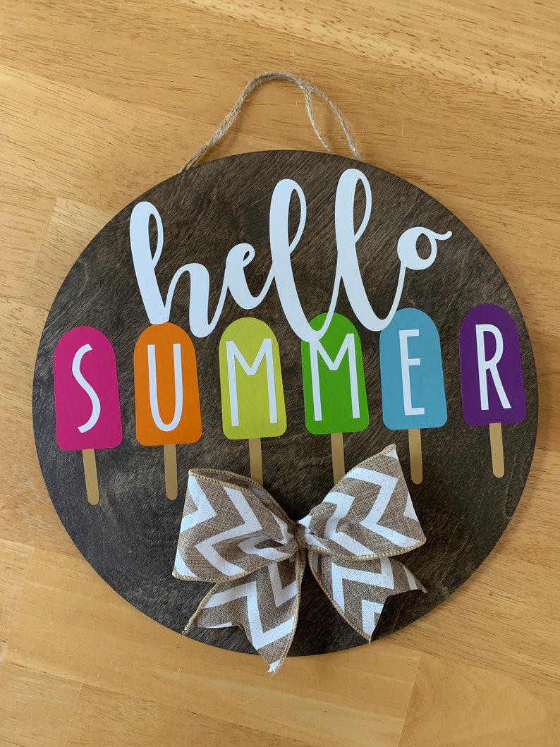 May include: A round wooden sign with the words "hello summer" in white script. The word "summer" is spelled out with colorful popsicle-shaped letters. A brown and white chevron bow is tied at the bottom of the sign.