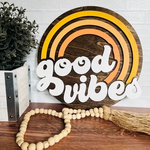 May include: A wooden round sign with a rainbow design in shades of yellow and orange. The sign says "good vibes" in white letters.