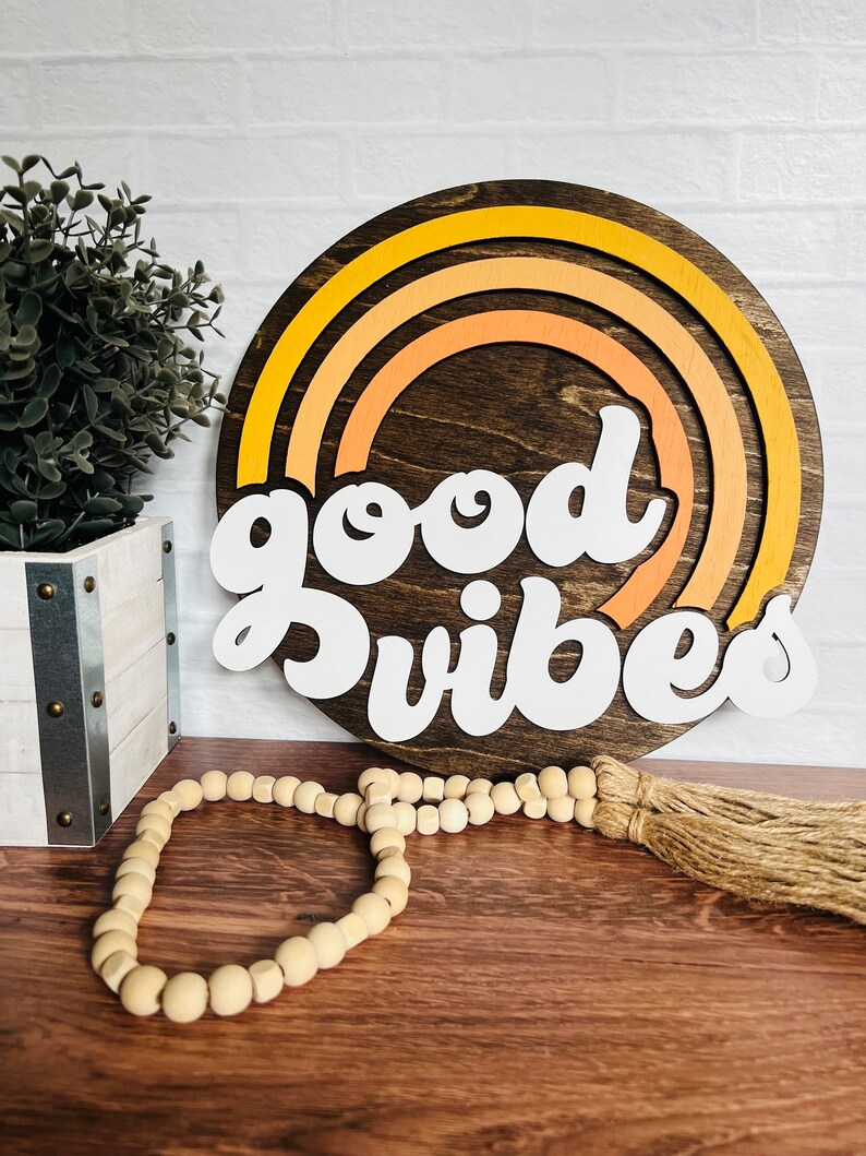 May include: A wooden round sign with a rainbow design in shades of yellow and orange. The sign says "good vibes" in white letters.