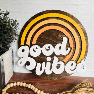 May include: A round wooden sign with a rainbow design in shades of orange and yellow. The sign says "good vibes" in white letters.