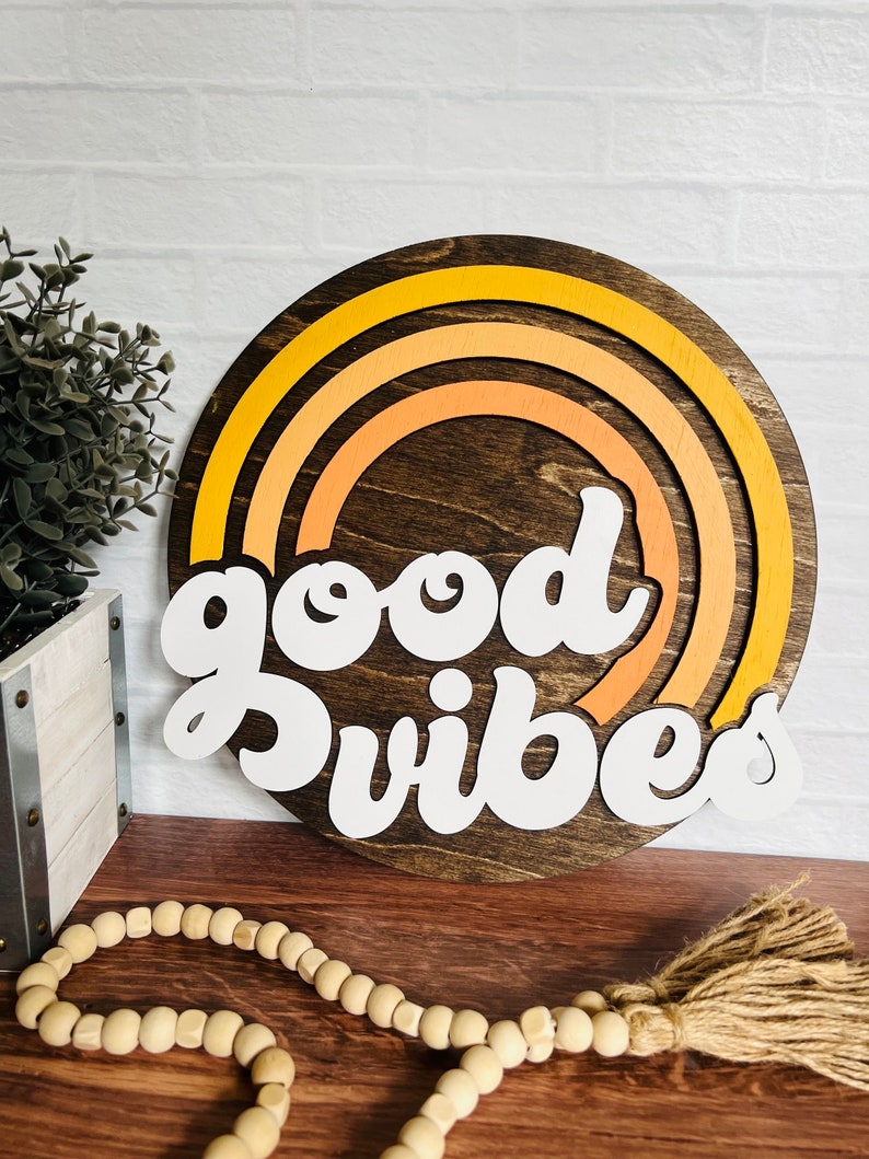 May include: A round wooden sign with a rainbow design in shades of orange and yellow. The sign says "good vibes" in white letters.