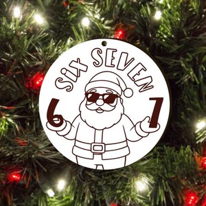 May include: A white circular Christmas ornament featuring a Santa Claus graphic wearing sunglasses and holding the numbers 6 and 7. The words "SIX SEVEN" are above the Santa illustration. The ornament has a hole for hanging.