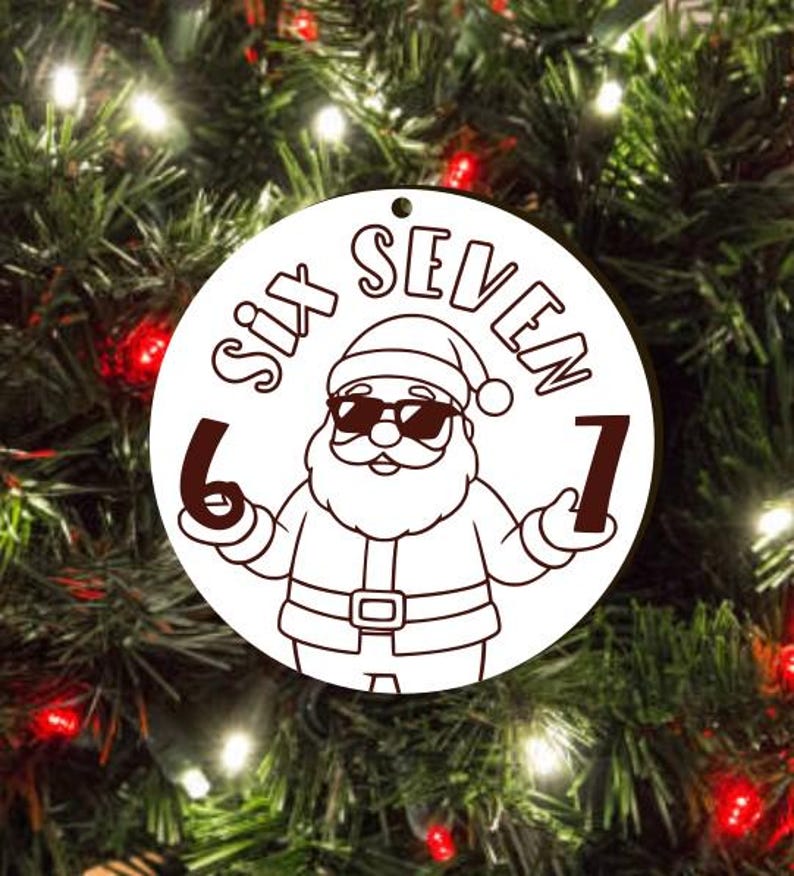 May include: A white circular Christmas ornament featuring a Santa Claus graphic wearing sunglasses and holding the numbers 6 and 7. The words "SIX SEVEN" are above the Santa illustration. The ornament has a hole for hanging.