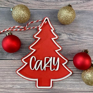 K&ouml;nnte beinhalten: Roter Weihnachtsbaum-Anh&auml;nger aus Holz mit wei&szlig;em Rand und dem Namen "Carly" in wei&szlig;. Der Anh&auml;nger ist von goldenen und roten Kugeln umgeben.