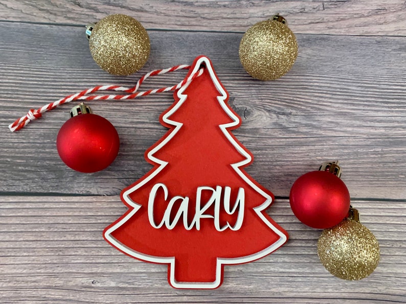 K&ouml;nnte beinhalten: Roter Weihnachtsbaum-Anh&auml;nger aus Holz mit wei&szlig;em Rand und dem Namen "Carly" in wei&szlig;. Der Anh&auml;nger ist von goldenen und roten Kugeln umgeben.