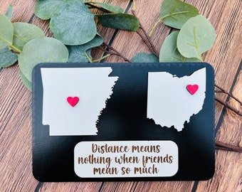 May include: A black wooden sign with white outlines of the states of Arkansas and Ohio. The states have red hearts on them. The sign says "Distance means nothing when friends mean so much."