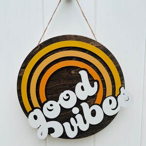 May include: A round wooden sign with a rainbow design in shades of yellow and orange. The sign says "good vibes" in white lettering.