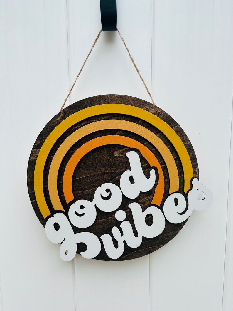 May include: A round wooden sign with a rainbow design in shades of yellow and orange. The sign says "good vibes" in white lettering.