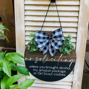Puede incluir: Un letrero redondo de madera con un lazo a cuadros blanco y negro y vegetaci&oacute;n. El letrero dice "no soliciting unless you brought alcohol, my amazon package, or Girl Scout cookies."