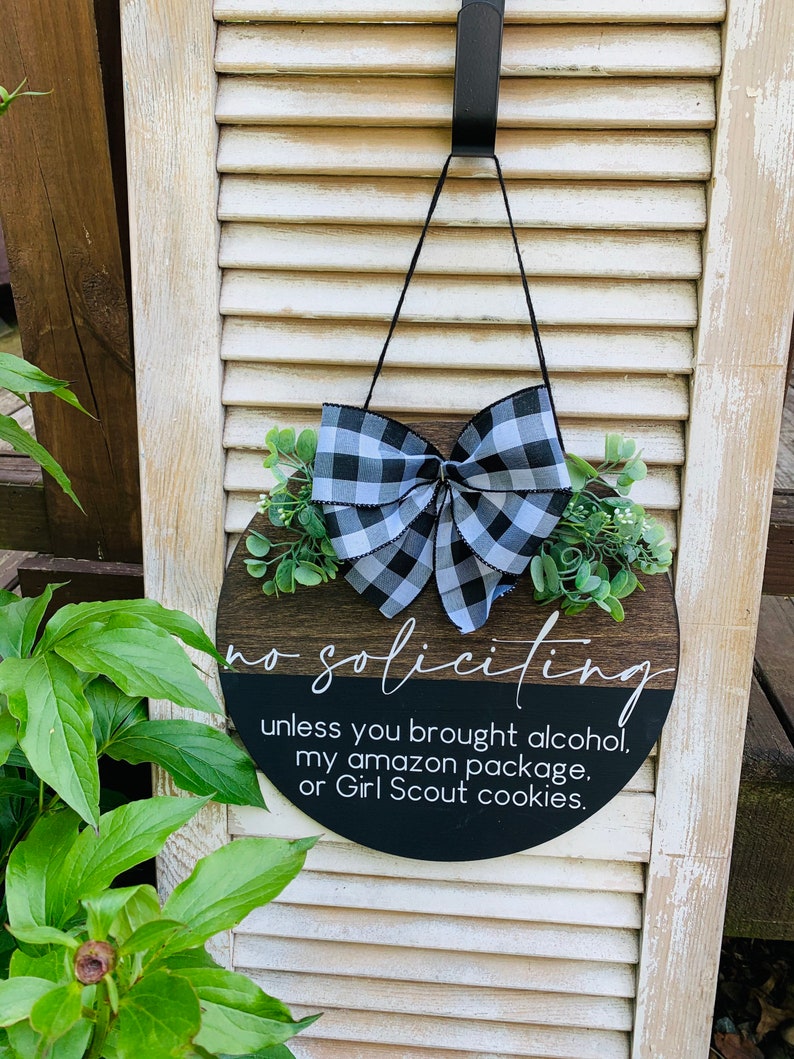 Puede incluir: Un letrero redondo de madera con un lazo a cuadros blanco y negro y vegetaci&oacute;n. El letrero dice "no soliciting unless you brought alcohol, my amazon package, or Girl Scout cookies."