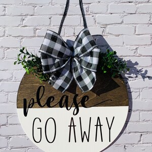 May include: A round wooden door hanger with a black and white checkered bow, green foliage, and the text "Please Go Away" in black lettering.